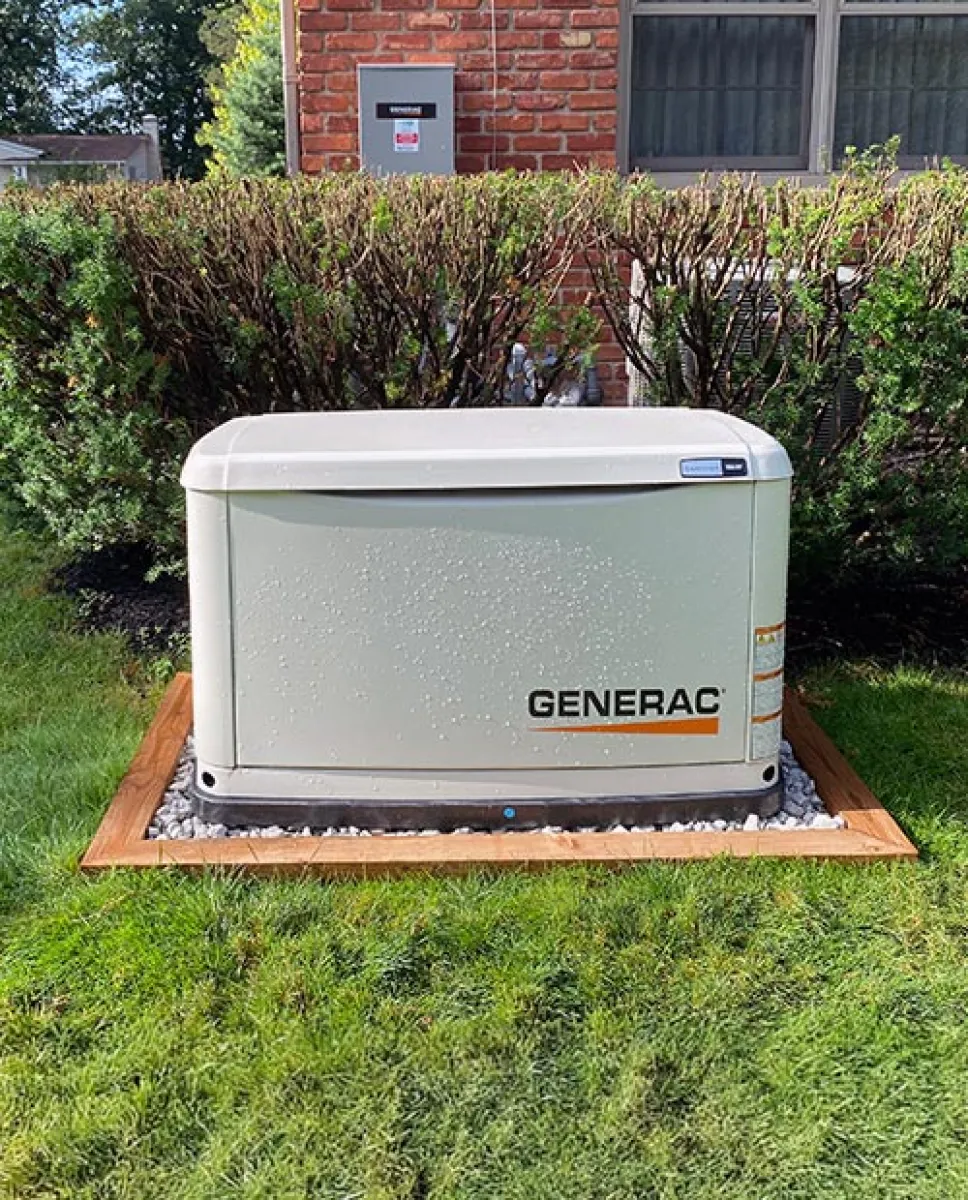 Generac home standby generator installed on grass next to brick house and trimmed bushes on a sunny day.