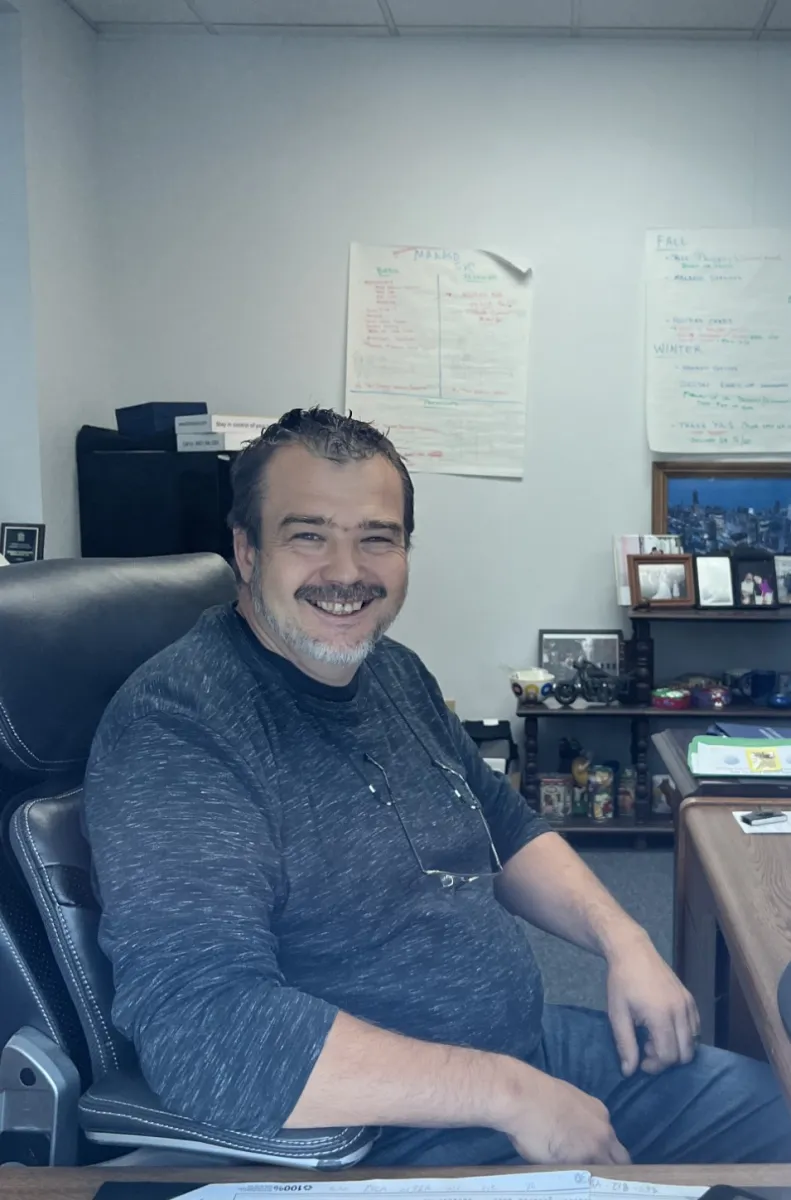 Smiling man with gray beard sitting in office chair at desk with computer, calendar, and office supplies.
