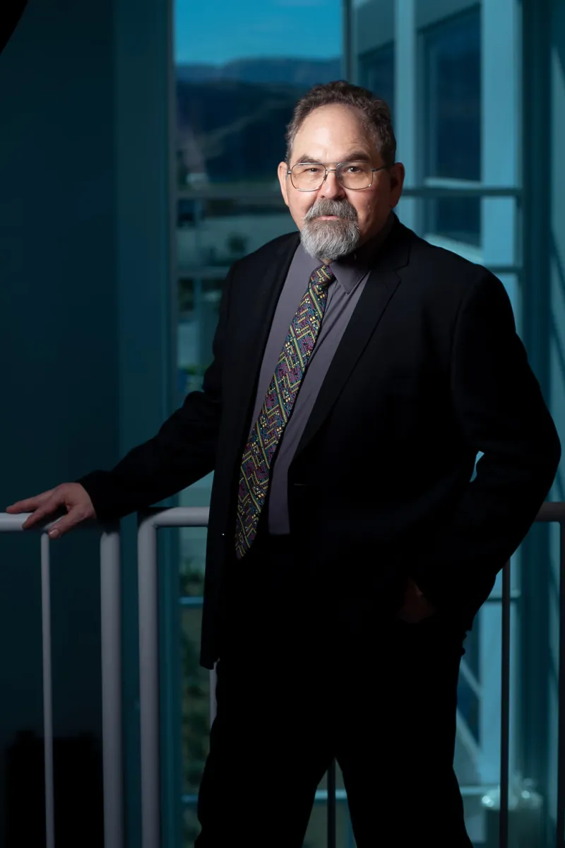 Middle-aged man with glasses and beard wearing a dark suit and colorful tie, standing indoors by a window.