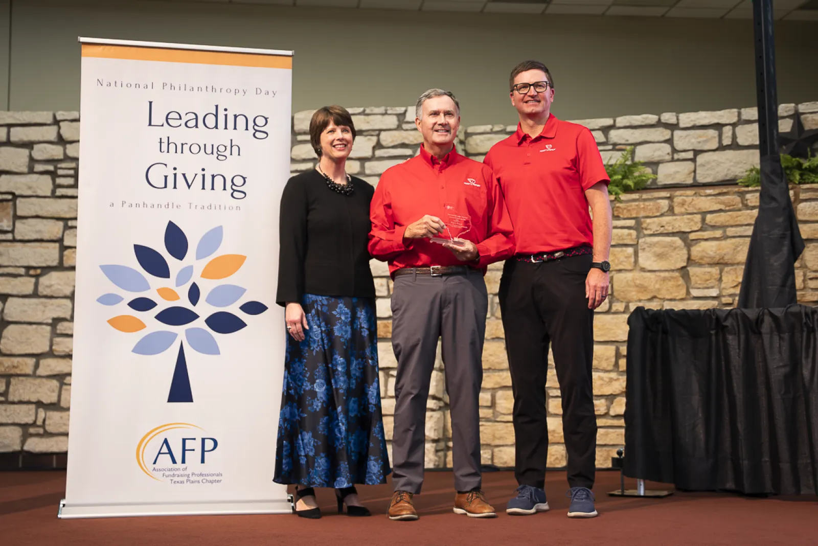 Three people stand next to AFP banner at National Philanthropy Day event, two men in red shirts holding an award.