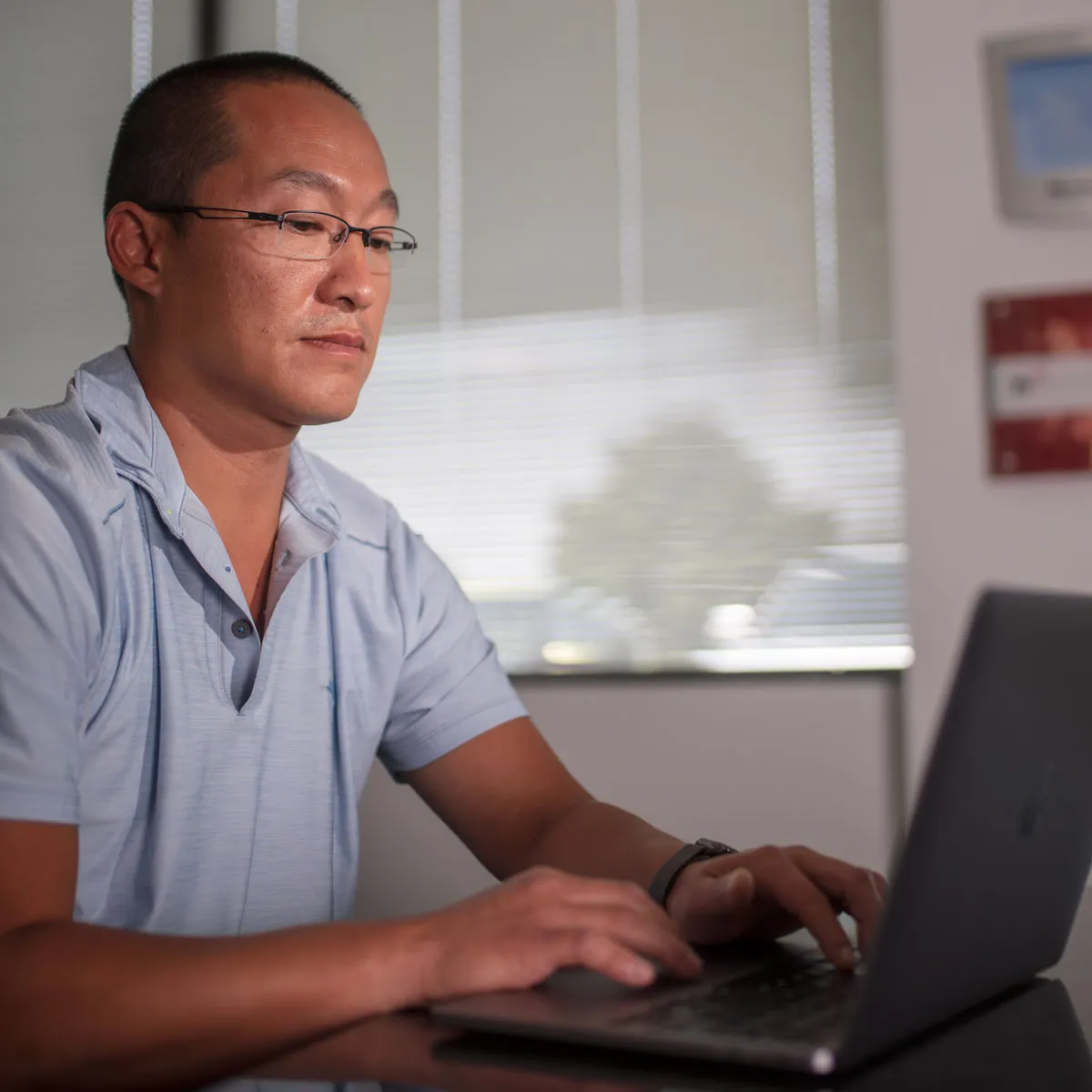 Man wearing glasses working on a laptop in a bright office with blinds and framed pictures.