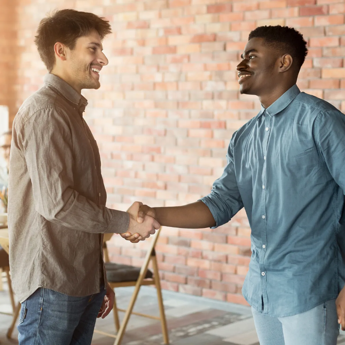 Two men smiling and shaking hands indoors with a brick wall background, symbolizing agreement or partnership