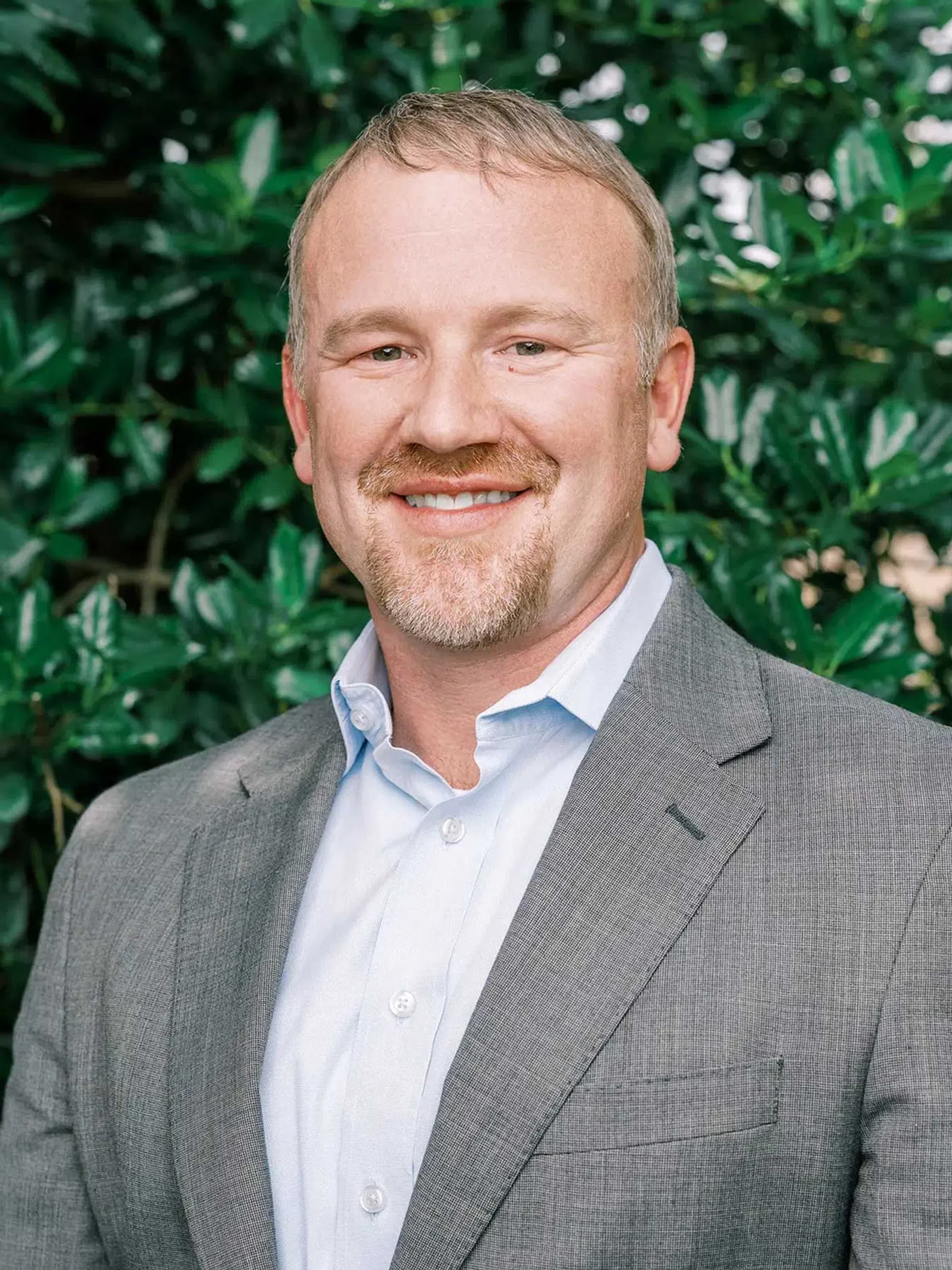 Smiling middle-aged man with short hair and beard wearing a light blue shirt and gray blazer against green foliage background