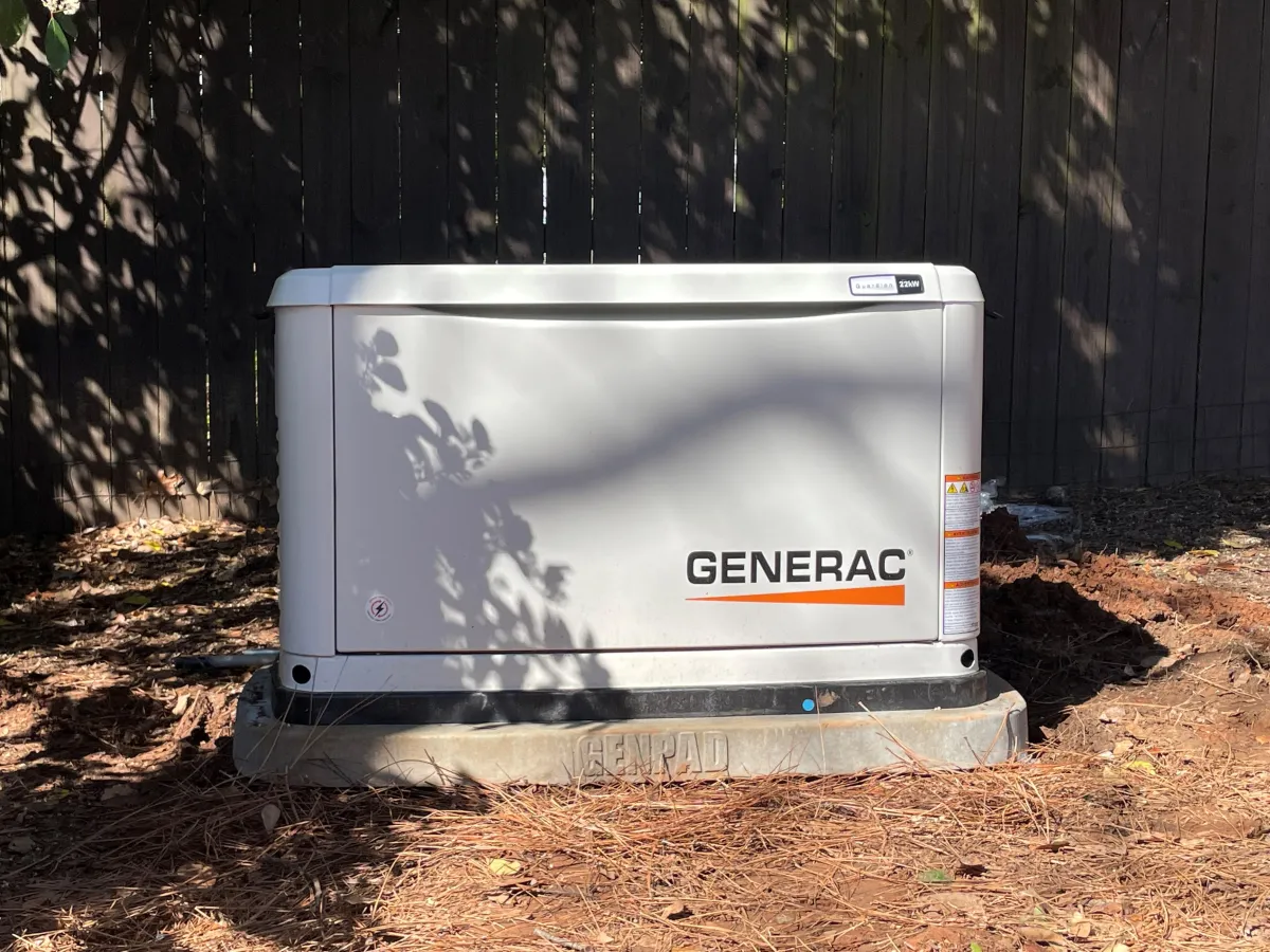 Generac home generator in a yard with sunlight filtering through trees and pine needles.