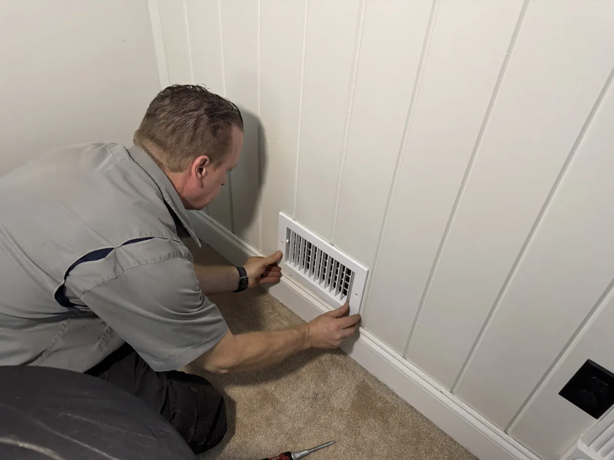 Technician installing a white wall AC vent cover on a beige carpeted floor against a white paneled wall.