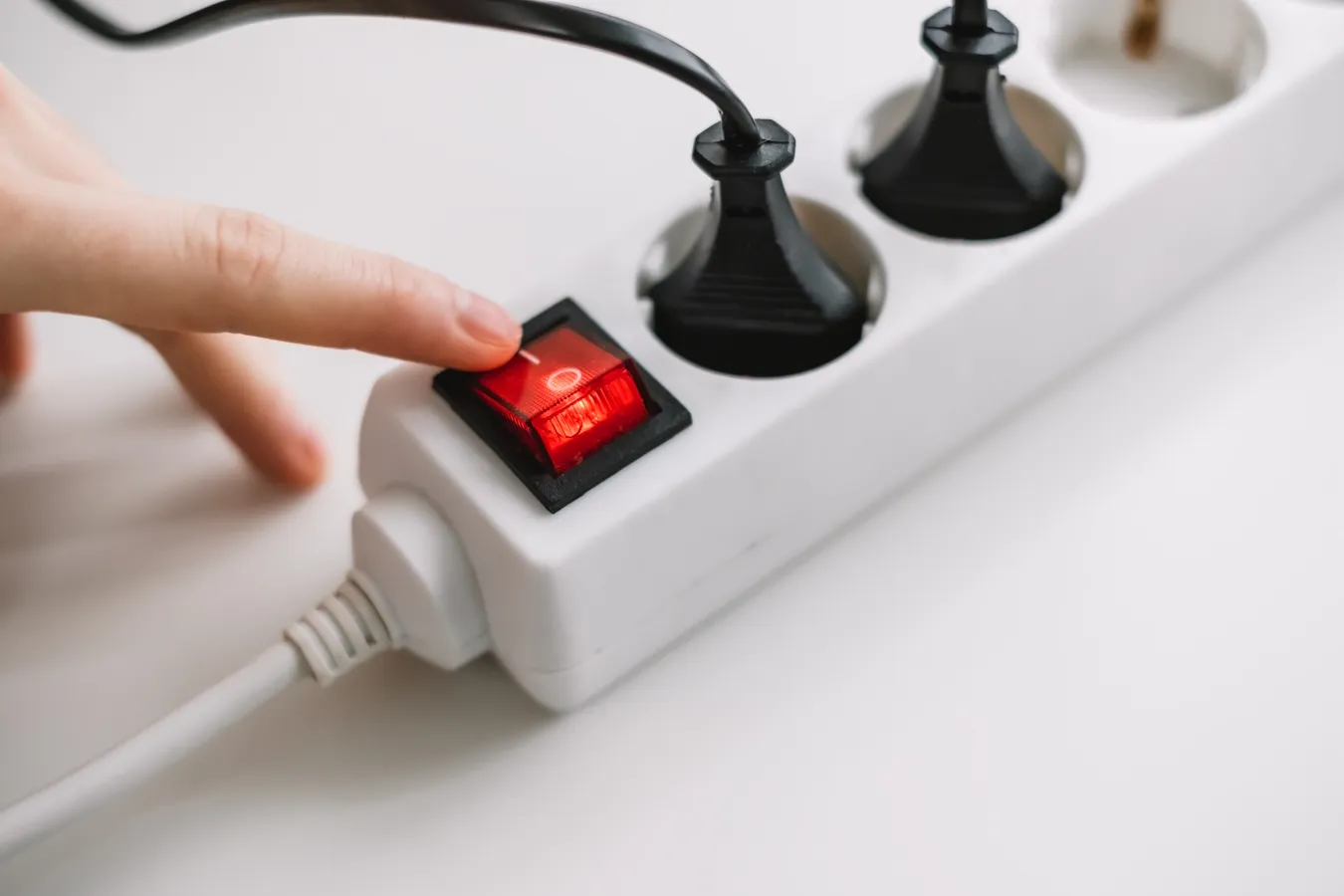 A hand pressing the switch on a white power strip with red indicator light.