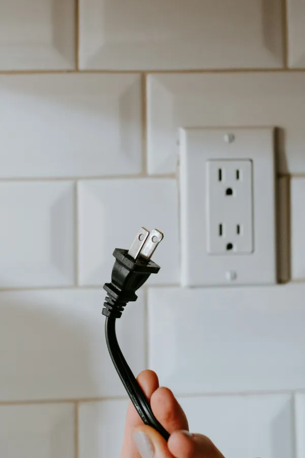 A close-up of a hand holding a power plug next to an electrical outlet on tiled wall.