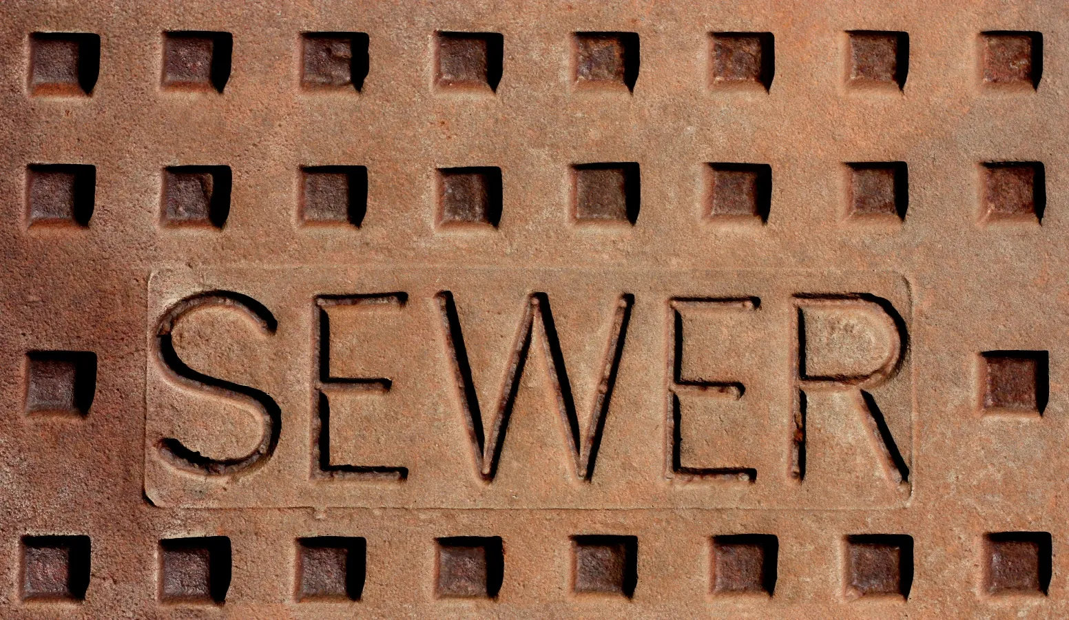 Close-up of a rusty metal sewer cover with the word SEWER engraved and square indentations.