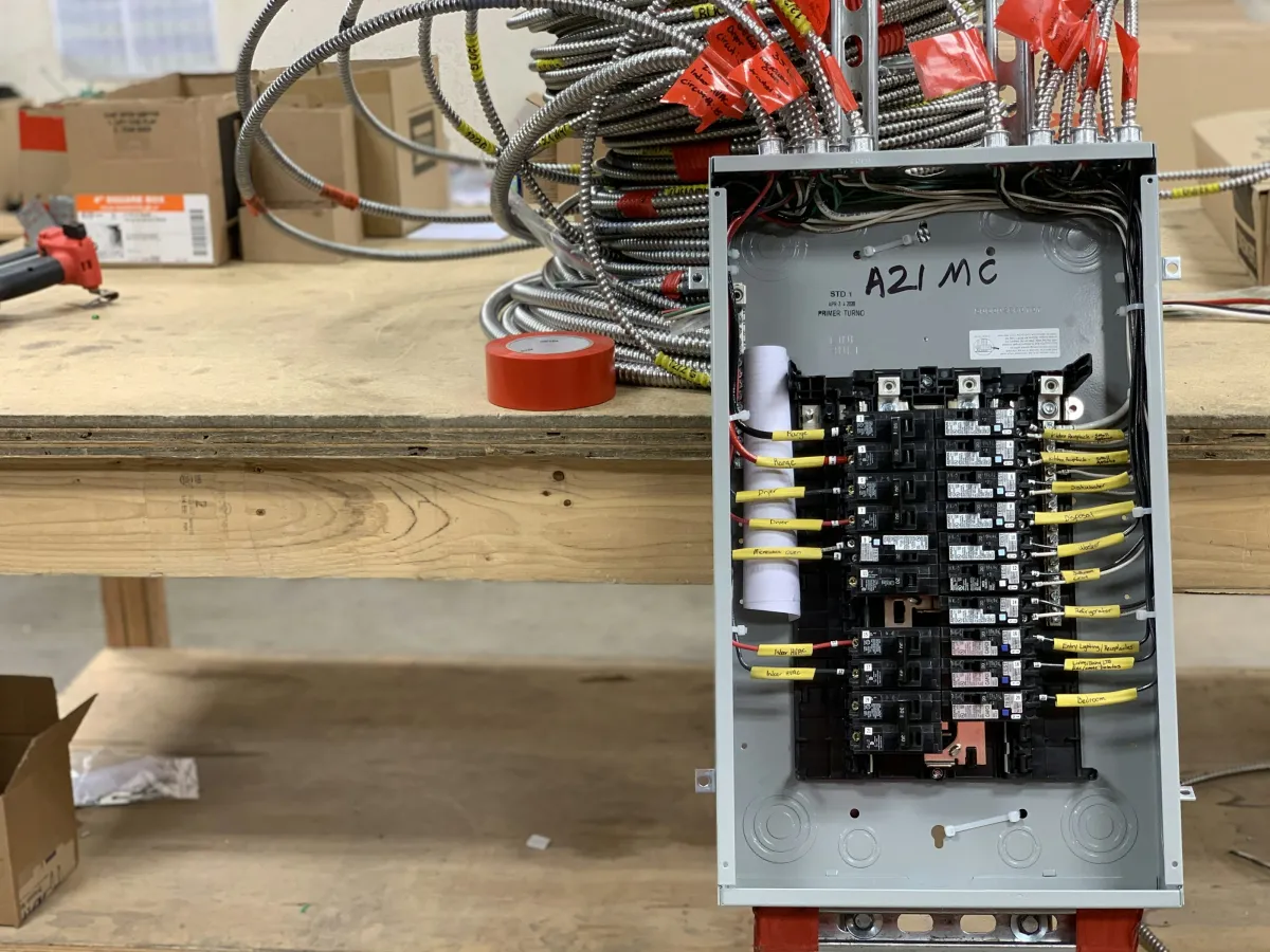 Open electrical breaker panel with labeled yellow wires on a wooden workbench with tools and cables.