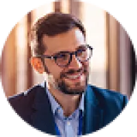 Smiling man with glasses and beard wearing a suit jacket in a warmly lit indoor setting