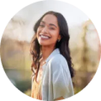 Smiling young woman with dark hair outdoors in soft sunlight wearing a light-colored top and jacket.