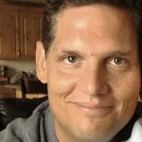 Close-up of a smiling man with short brown hair in a home setting with wooden cabinets in the background