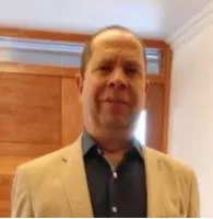 Man wearing beige blazer and dark shirt standing indoors with wooden door and window in background.