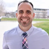 Smiling man wearing a light purple shirt and colorful plaid tie outdoors with blurred buildings and grass background