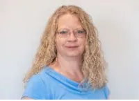 Portrait of a woman with curly blonde hair wearing glasses and a blue top against a plain light background
