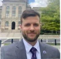 Man with a beard in suit and tie standing outdoors near a historic building and greenery on a sunny day