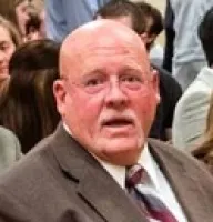 Elderly man in a brown suit and striped tie in a crowded indoor setting with people in the background