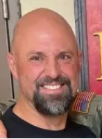 Smiling bald man with a dark beard and mustache, wearing a black shirt, posing indoors.