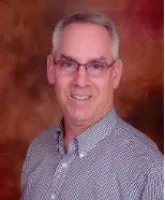 Middle-aged man with gray hair and glasses smiling against a warm brown backdrop, wearing a patterned button-up shirt