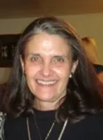 Smiling middle-aged woman with long brown hair wearing a black top and pearl earrings indoors.