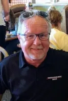 Older man with glasses and sunglasses on head smiling in a busy restaurant setting wearing a black polo shirt.