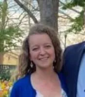 Smiling woman with curly hair wearing a blue cardigan outdoors in a garden setting with trees and flowers.