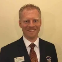 Smiling man in black suit, white shirt, and patterned tie against a beige background.