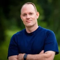Smiling man in a blue shirt with arms crossed standing outdoors with a blurred green background
