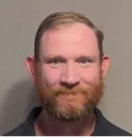 Close-up of a man with a red beard, short hair, and blue eyes against a plain gray background.