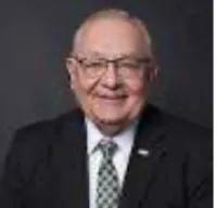 Elderly man wearing glasses and a suit with a patterned tie smiling against a dark background.