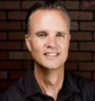 Portrait of a middle-aged man smiling in front of a dark brick wall background.