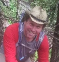 Smiling man wearing a cowboy hat and red jacket hiking in a forested area