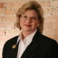 Professional woman with glasses and short blonde hair wearing black blazer and white shirt against brick wall.