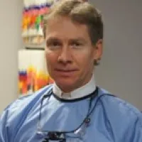 Male dentist wearing a blue dental gown and magnifying glasses standing in a clinic with colorful charts in background
