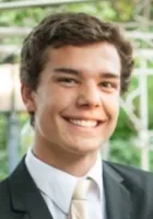 Young man smiling in a suit and tie with a green outdoor background and a white fence.