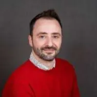Smiling man with short dark hair and beard wearing a red sweater over a patterned shirt on gray background