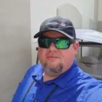 Man wearing reflective sunglasses, black cap, and blue shirt standing outdoors near a golf cart.