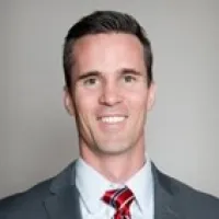 Professional headshot of a smiling man in a grey suit and red striped tie against a neutral background