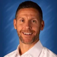 Smiling man with short hair and beard wearing a white shirt against a blue studio background