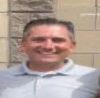 Smiling man in light gray polo shirt standing outside against a beige and brick wall background.