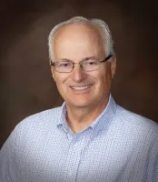 Smiling older man with glasses wearing a light blue checkered shirt against a brown background.