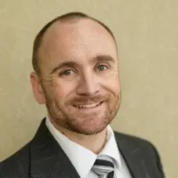 Smiling bald man with beard wearing a dark suit, white shirt, and striped tie against a beige background