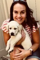 Smiling woman in striped shirt holding a white Labrador puppy outdoors near a fence.