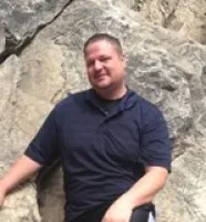 Man wearing a dark polo shirt sitting and smiling against a rocky background outdoors