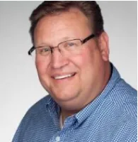 Smiling man wearing glasses and a blue checkered shirt against a plain light background.