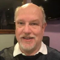 Smiling middle-aged man with a white beard and balding head wearing a collared shirt and sweater indoors.