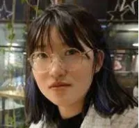Young woman with glasses and dark hair sitting indoors in a casual setting with plants in the background