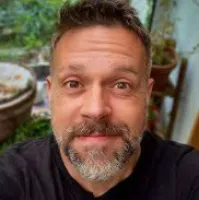 Close-up of a middle-aged man with a beard smiling indoors with blurred plants and furniture in the background
