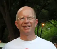 Smiling man with glasses wearing a white t-shirt standing outdoors near trees and street lamps at dusk