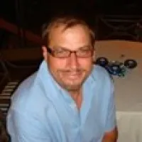 Man wearing glasses and a light blue shirt sitting at a table with a floral centerpiece indoors.